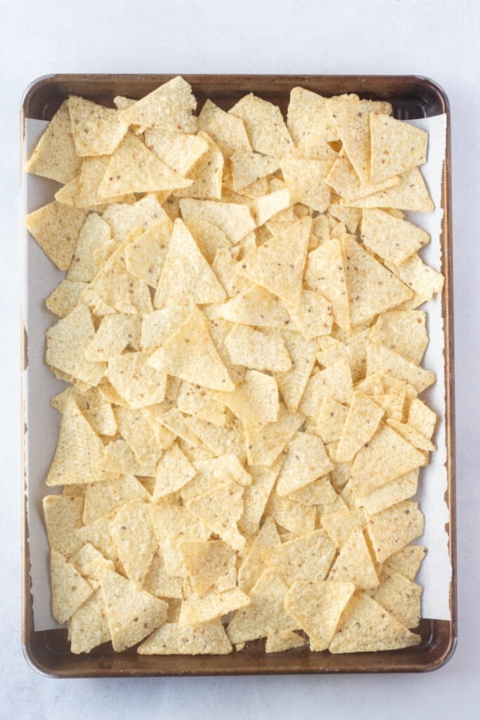 A top down shot of a sheet pan lined with white parchment paper that also has a layer of white corn tortilla chips on it.