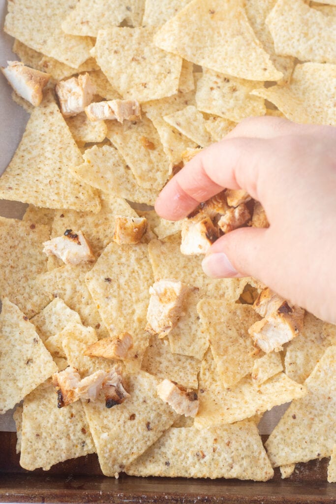 A hand placing pieces of cooked chicken on top of a layer of white corn tortilla chips that are on a sheet pan lined with parchment paper.