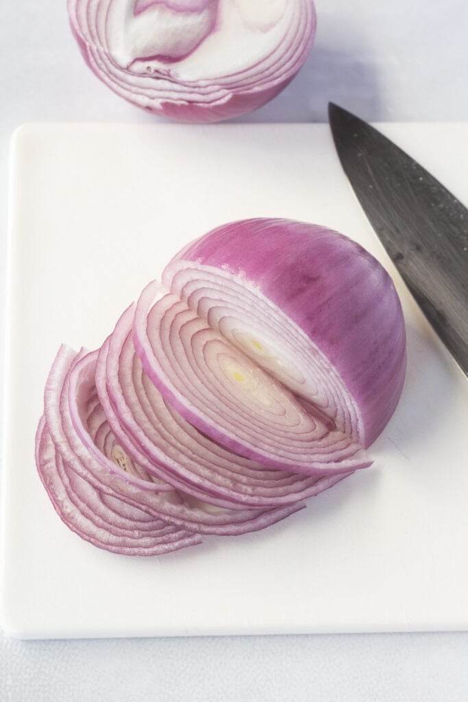 Half a red onion that's been partially sliced on a white cutting board.