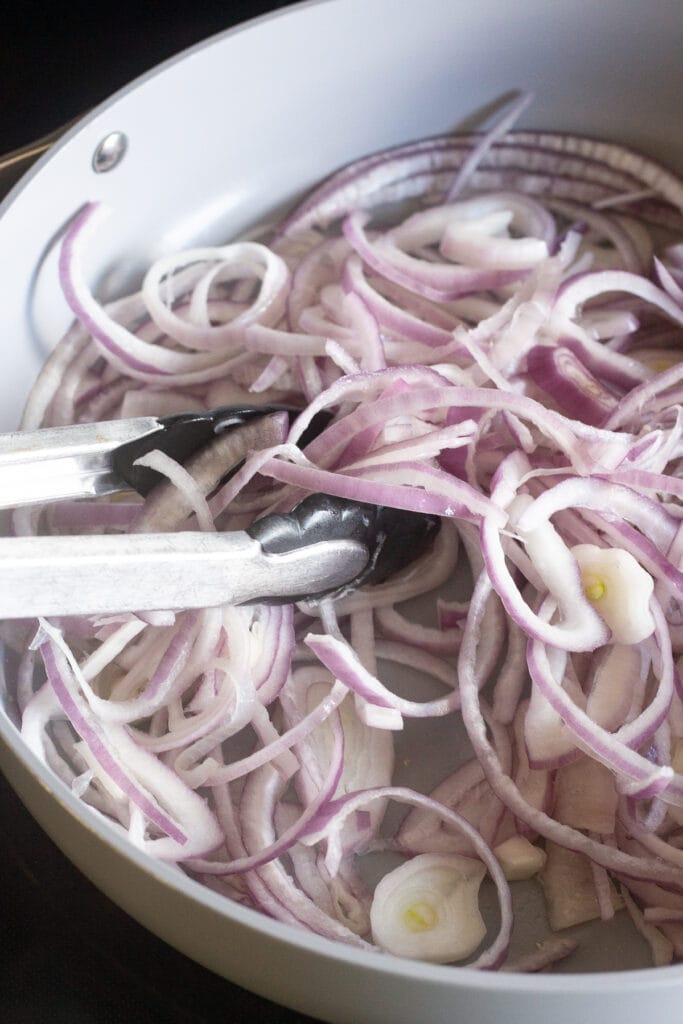 Sliced purple onions being tossed with a pair of tongs in a large gray pan on a stovetop.