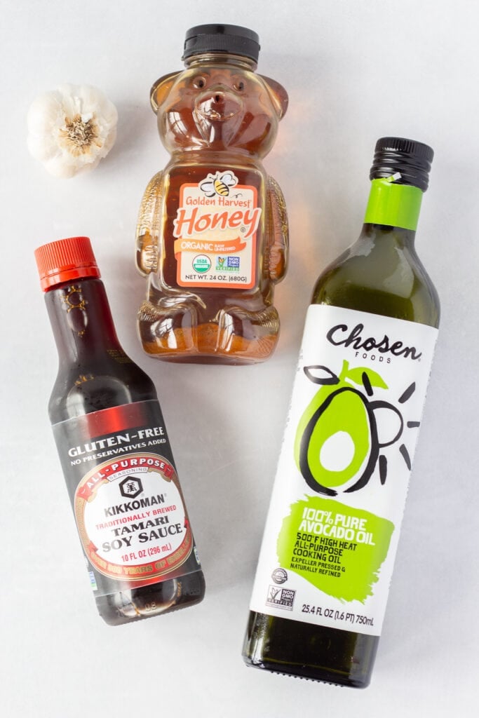 Top down shot of a bottle of gluten free soy sauce, a bottle of avocado oil, a bear-shaped jar of honey, and a head of garlic on a white background.