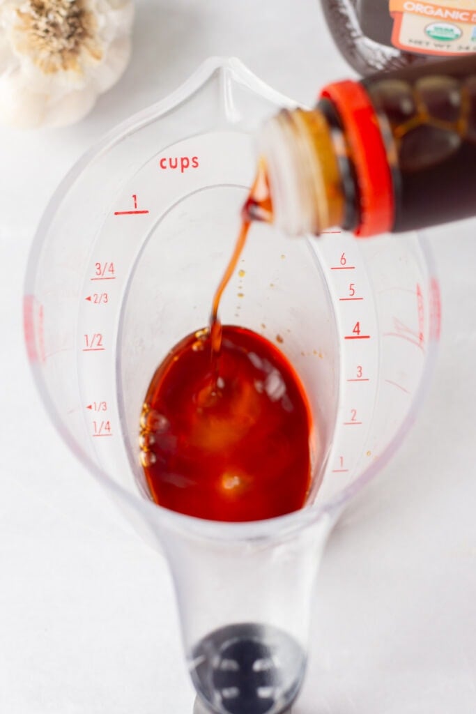 Soy sauce being pouring into a clear plastic measuring cup on a white surface.