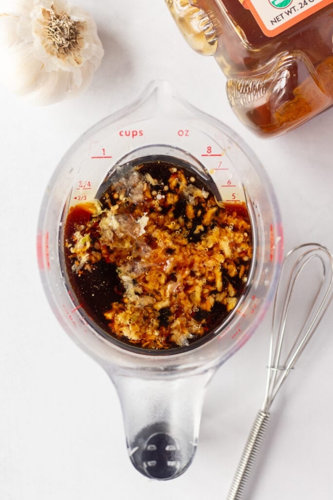 Top down shot of a measuring cup with soy sauce and garlic in it, on a white surface. Next to it are a head of garlic, a bear shaped bottle of honey, and a small whisk.