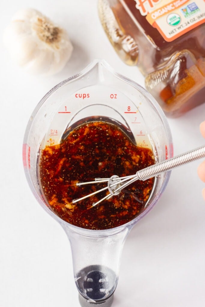 A top down shot of a small whisk stirring a garlic soy sauce marinade in a small clear plastic measuring cup with a head of garlic and a bear-shaped bottle of honey in the background.