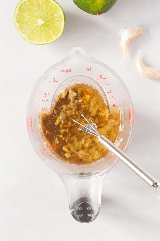 Top down shot of a brown marinade with garlic in it in a small measuring cup with a whisk sticking out.