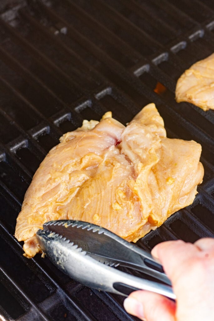 Metal tongs placing raw marinated chicken breasts on a hot grill.