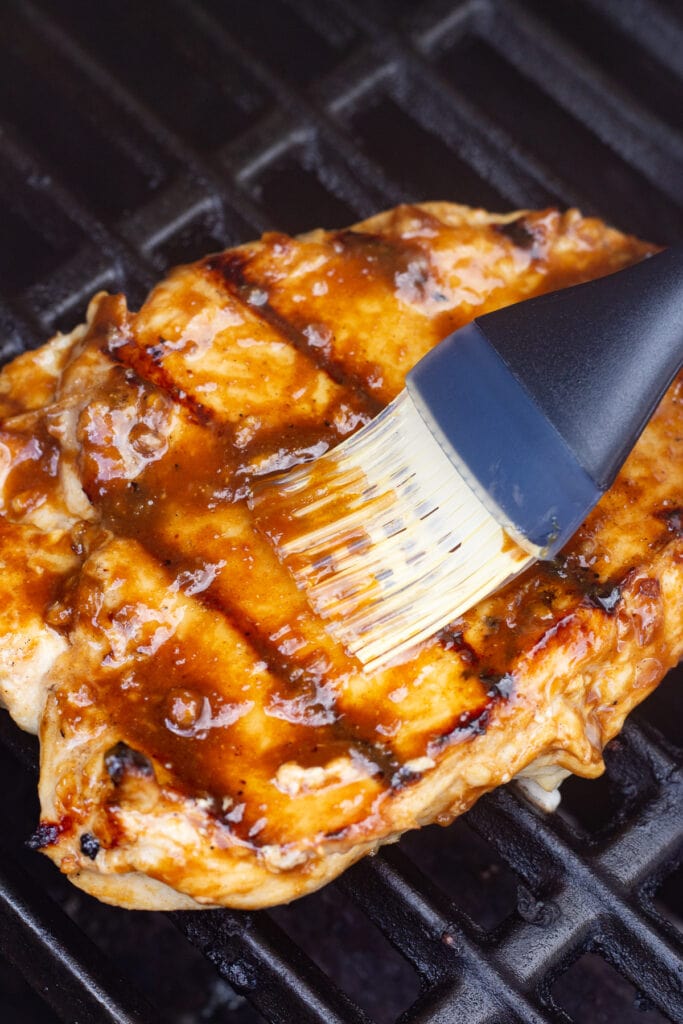 A basting brush spreading a brown glaze onto a grilled chicken breast that's on a hot grill.
