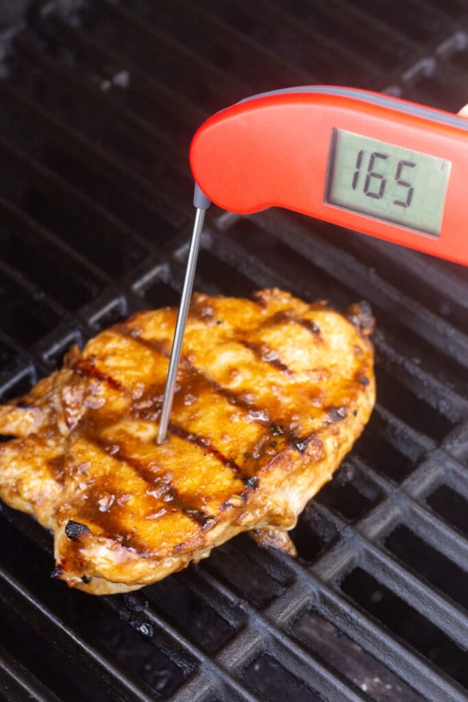 A red meat thermometer checking the temperature of a grilled chicken breast on the grill, measuring 165 degrees F.