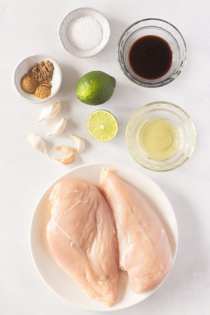 Top down shot of ingredients for grilled Mexican chicken breasts on a white background, including 2 raw chicken breasts on a white plate, 5 garlic cloves, one and a half limes, and bowls with spices and oils in them.