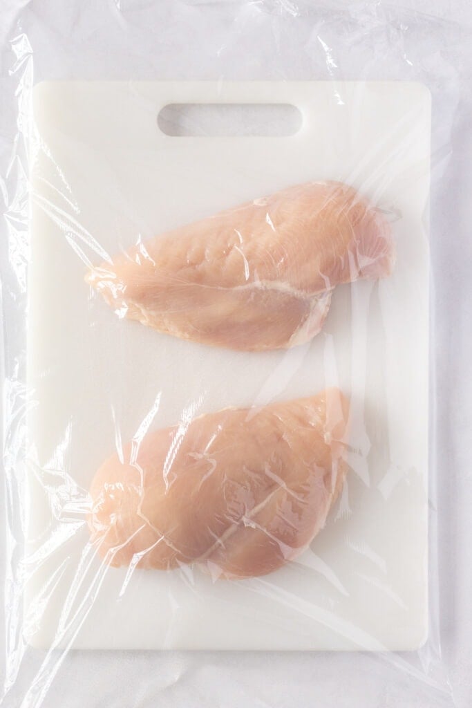 Top down shot of two raw chicken breasts on a white cutting board covered with plastic wrap.