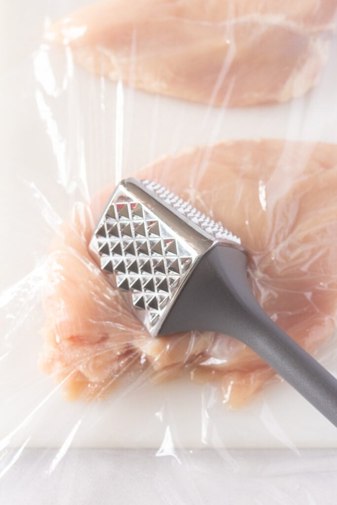 Close up of a meat mallet pounding a raw chicken breast on a white cutting board with a piece of plastic wrap on it.