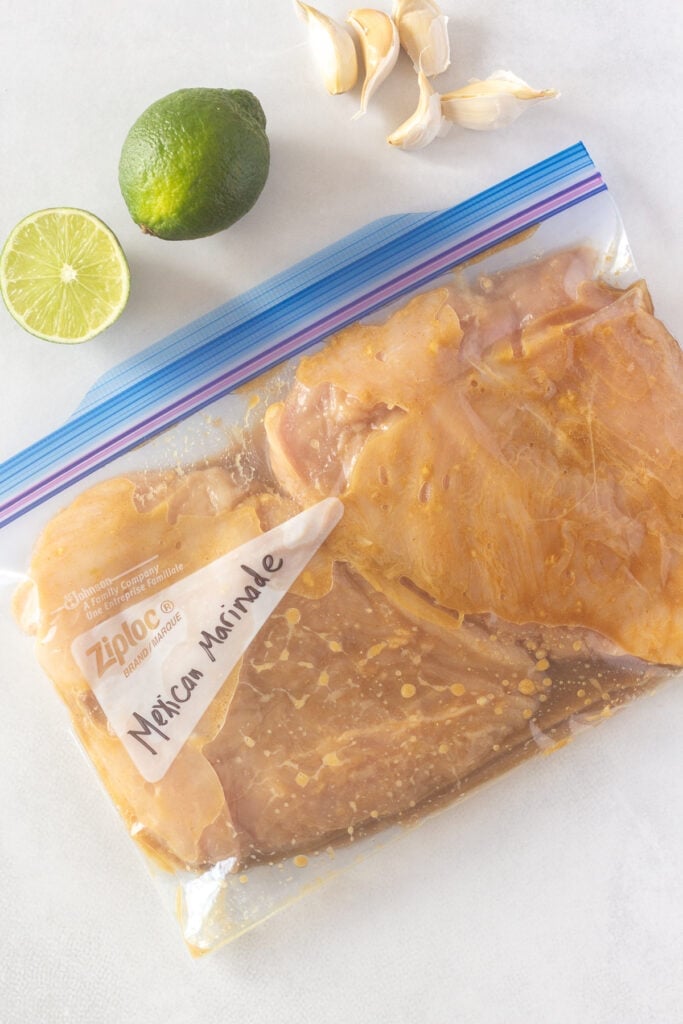 Top down shot of a ziploc bag with two flattened chicken breasts marinating in it. One and a half limes and five cloves of garlic are next to the bag.