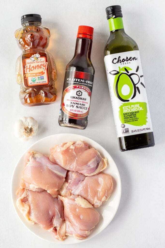 Top down shot of ingredients for grilled soy garlic chicken, including bottles of avocado oil, soy sauce, and honey, a clove of garlic, and a white plate with raw boneless skinless chicken thighs on it.