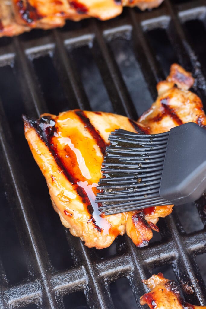 Close up of a black basting brush swiping a honey soy glaze on top of grilling boneless skinless chicken thighs on a grill.