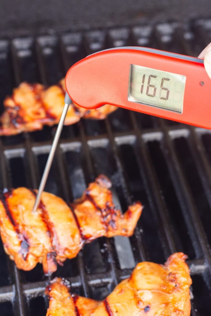 A red meat thermometer poking into grilled boneless skinless chicken thighs on a grill grate, showing their internal temperature at 165 degrees F.