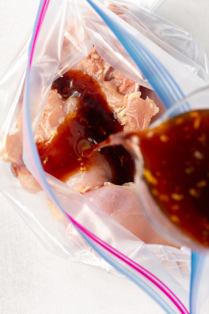 Close up of raw boneless skinless chicken thighs in a plastic ziploc bag with a soy sauce marinade being poured over them.
