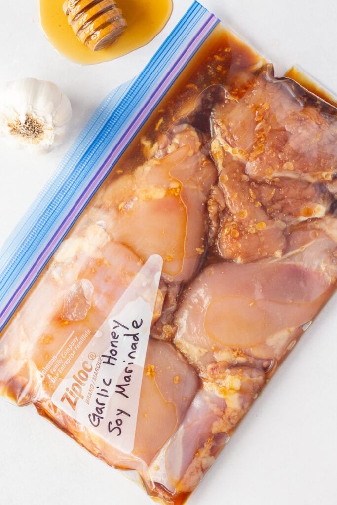 Top down shot of raw boneless skinless chicken thighs in a garlic honey soy marinade next to a head of garlic and some honey, all on a white background.