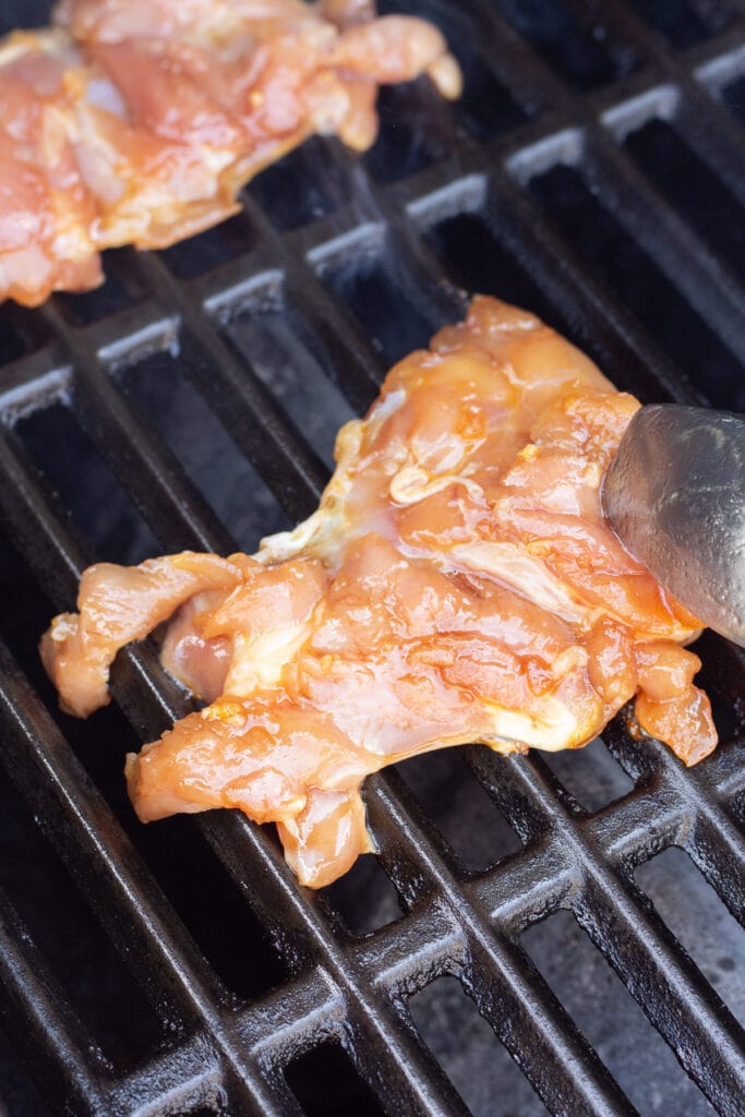 Close up of marinated raw boneless skinless chicken thighs being placed on a hot grill with tongs.