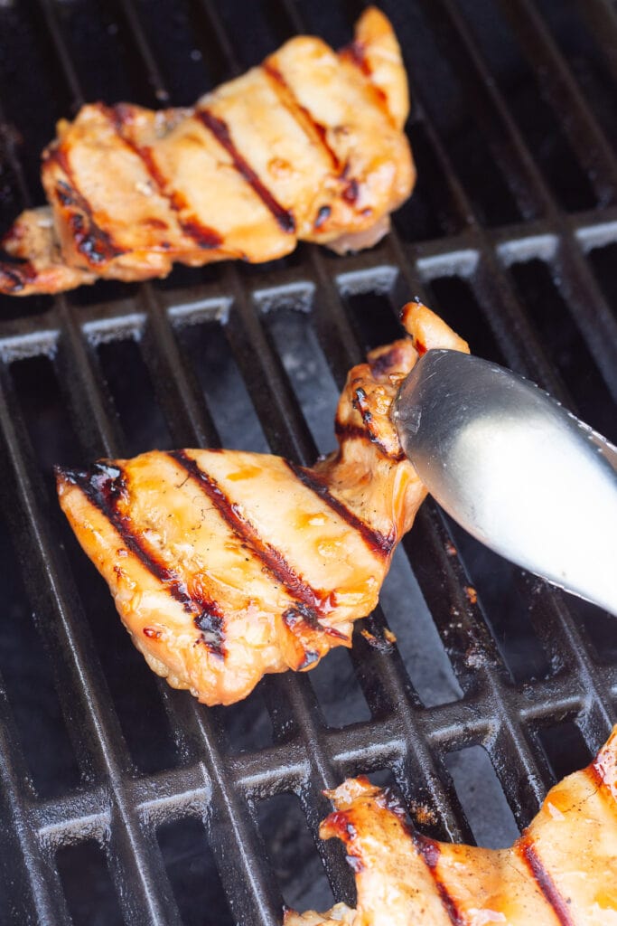 Close up of a pair of silver tongs flipping over boneless skinless chicken thighs on a grill.