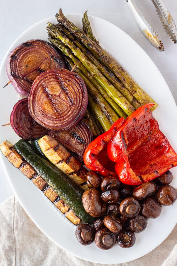 Top down shot of marinated and grilled red onion, zucchini, asparagus, mushrooms, and bell pepper on a white oval platter next to a pair of tongs and a tan napkin, all on a white background.