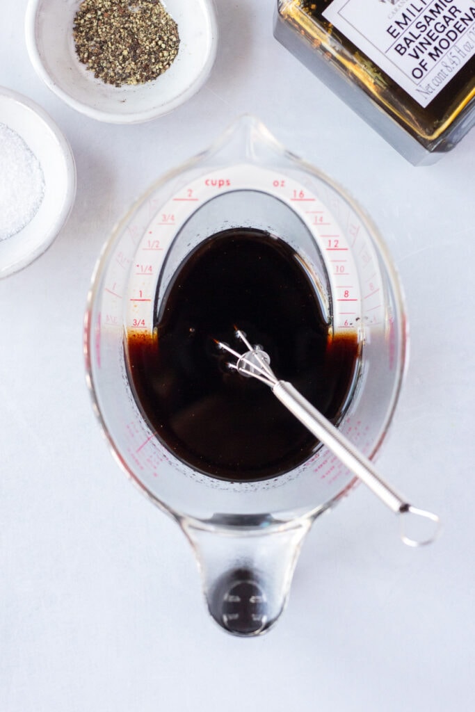 Top down shot of a plastic measuring cup with a balsamic marinade in it, with a whisk resting in it as well. Toward the top of the frame are two small white bowls with salt and pepper, and a bottle of balsamic vinegar, all on a white background.