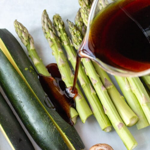 A measuring cup pouring a balsamic marinade over raw asparagus and zucchini in a glass dish.