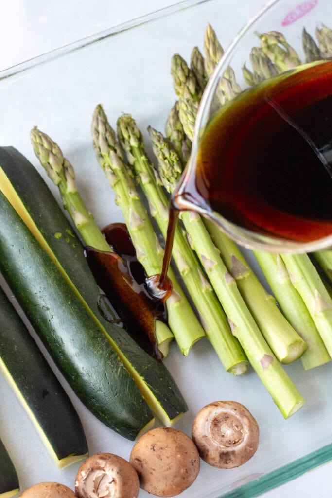 A measuring cup pouring a balsamic marinade over raw asparagus and zucchini in a glass dish.
