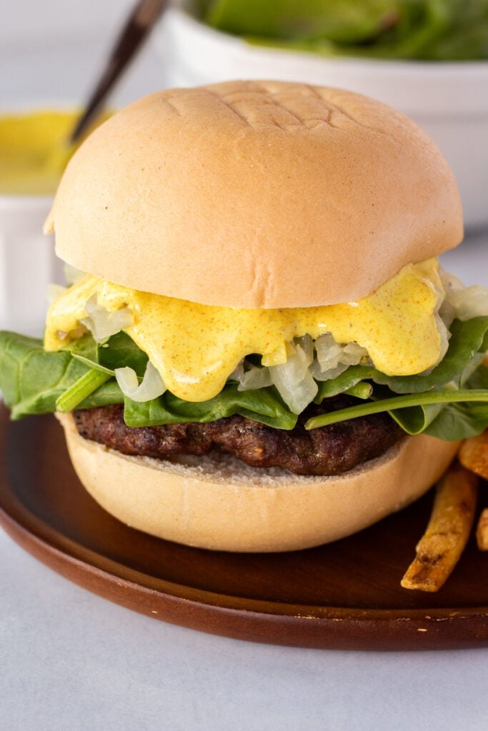 Close up of a curry burger on a gluten free bun topped with greens, sauteed onions, and a yellow curry sauce on a dark brown plate with fries to the side.