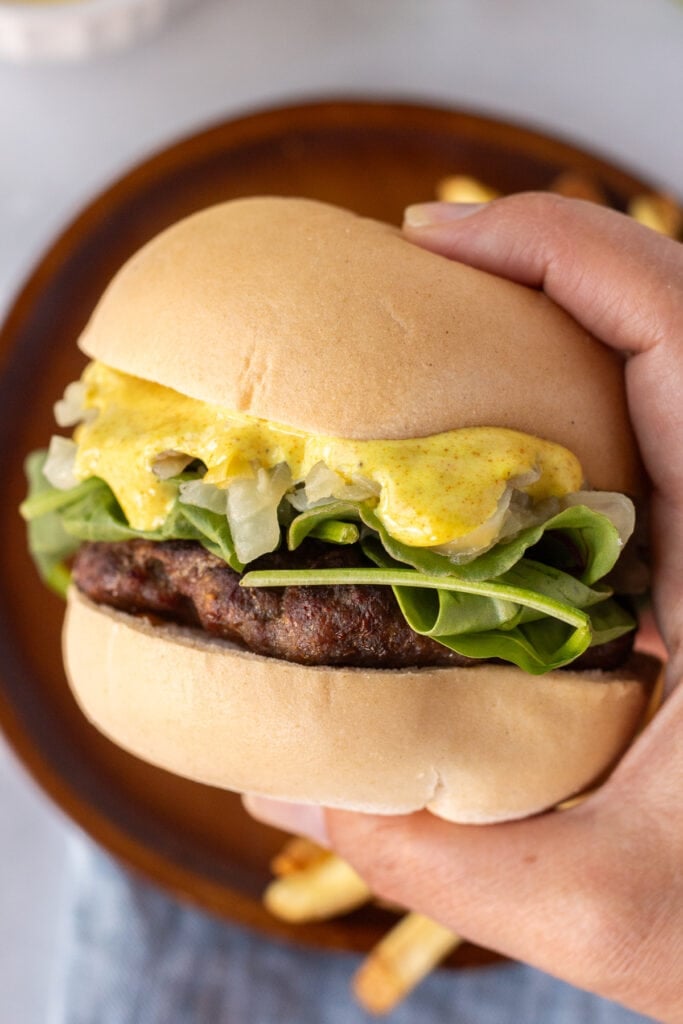 A hand holding a curry burger with a gluten free bun, some mixed greens, sauteed onions, and a yellow curry sauce.