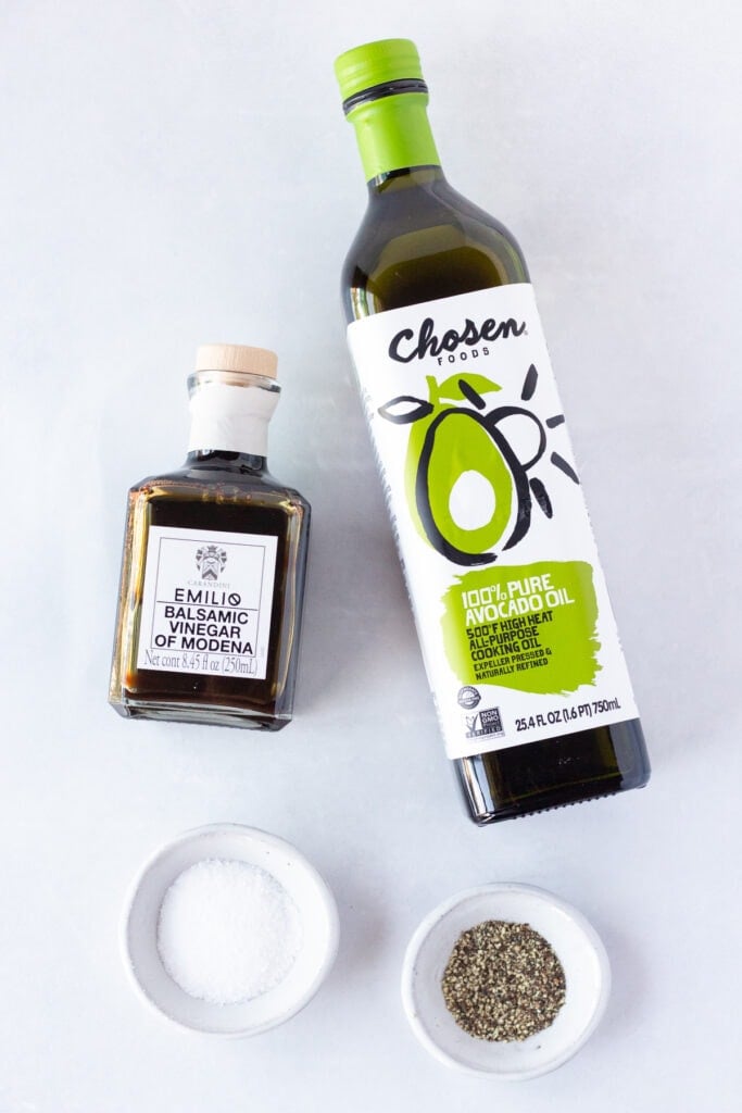 Top down shot of a bottle of avocado oil, a bottle of balsamic vinegar, and two small bowls with salt and pepper in them, respectively, all on a white background.