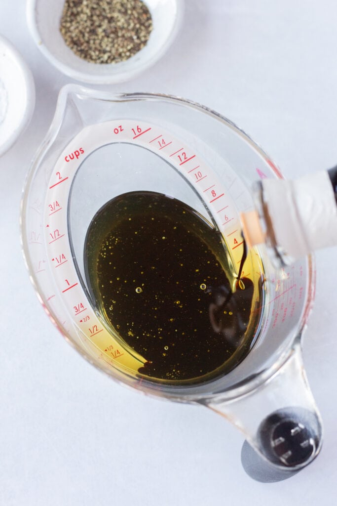 Pouring balsamic vinegar into a measuring cup with oil in it that's on top of a white background next to small bowls of salt and black pepper.