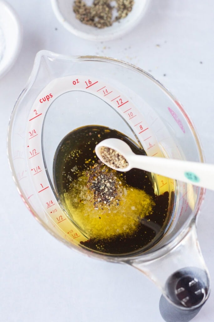 A small measuring spoon dumping ground black pepper into a measuring cup that has oil, vinegar, and salt already in it.