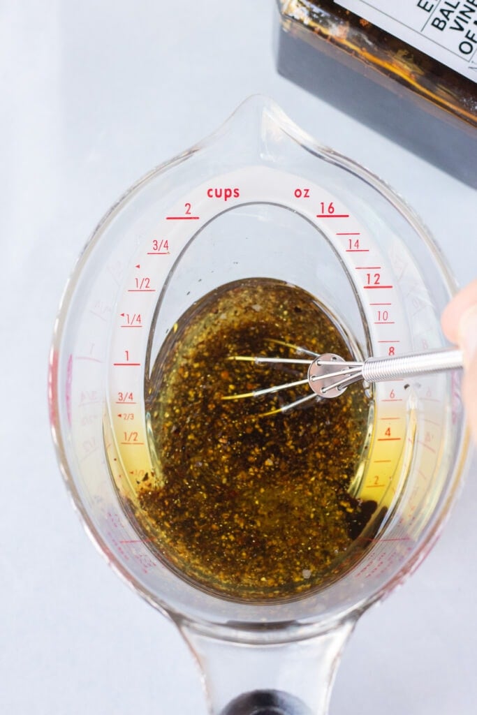 A hand using a small whisk to stir together vinegar, oil, salt, and pepper in a plastic measuring cup that's sitting on a white surface.