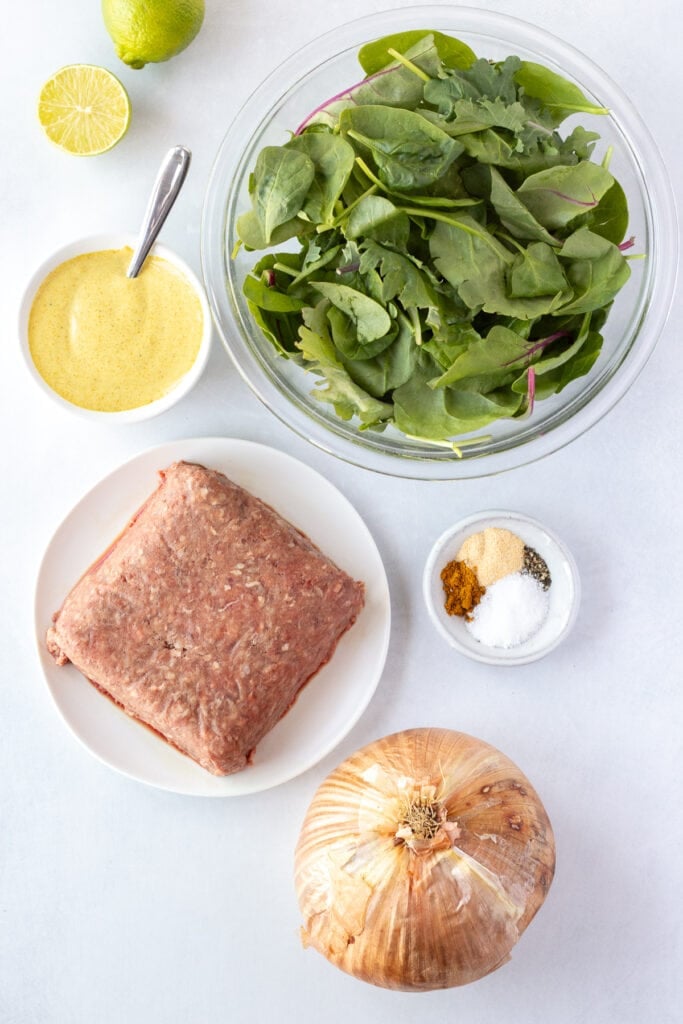 Top down shot of ingredients for curry burgers on a white backdrop, including a onion, a curry sauce, greens in a bowl, spices in a small bowl, and a white plate with a pound of raw ground beef.