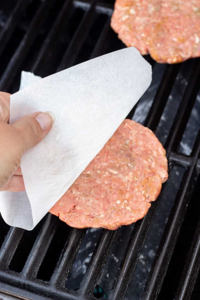Peeling white parchment paper off a raw burger patty after placing it on a hot grill.