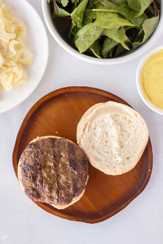 Top down shot of a curry burger on a gluten free bun that's on a dark wood plate. Next to the wood plate is a white plate with sauteed onions, a white bowl with mixed baby greens, and a small white bowl with yellow curry mayo sauce.