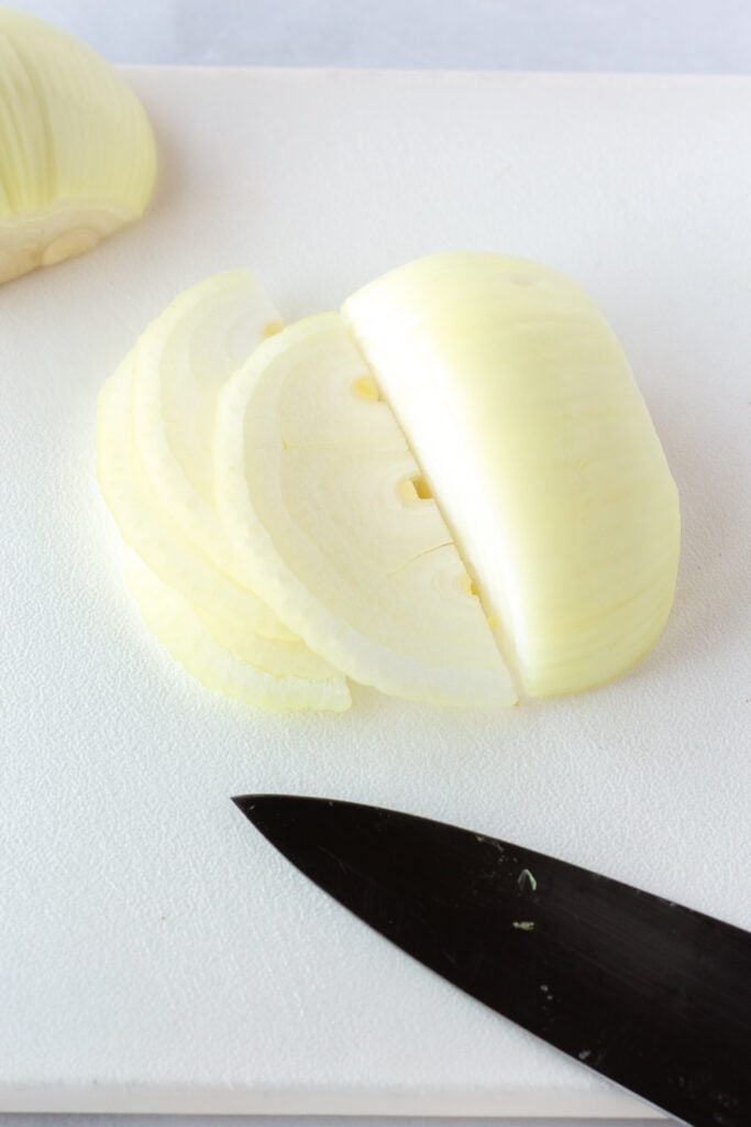 Close up of a white cutting board with an yellow onion being thinly sliced on it.
