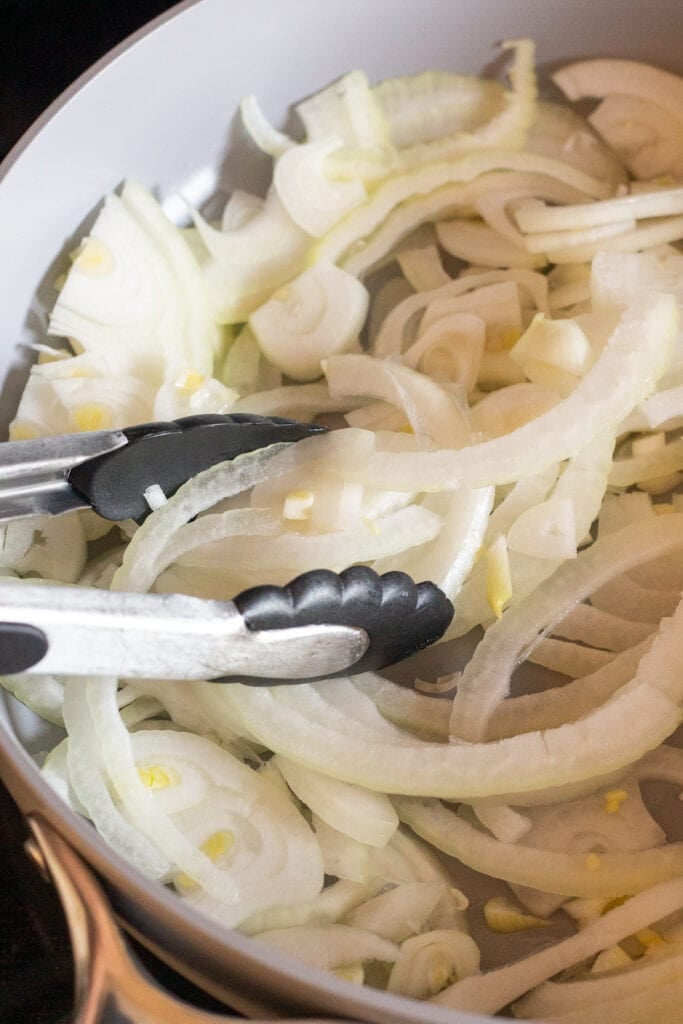 Close up of sliced yellow onions being sauteed in a large gray nonstick pan with tongs stirring them.
