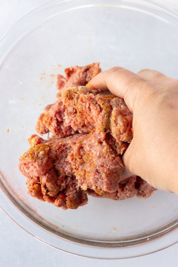 Close up of a hand smushing seasonings into some raw ground beef in a clear bowl.