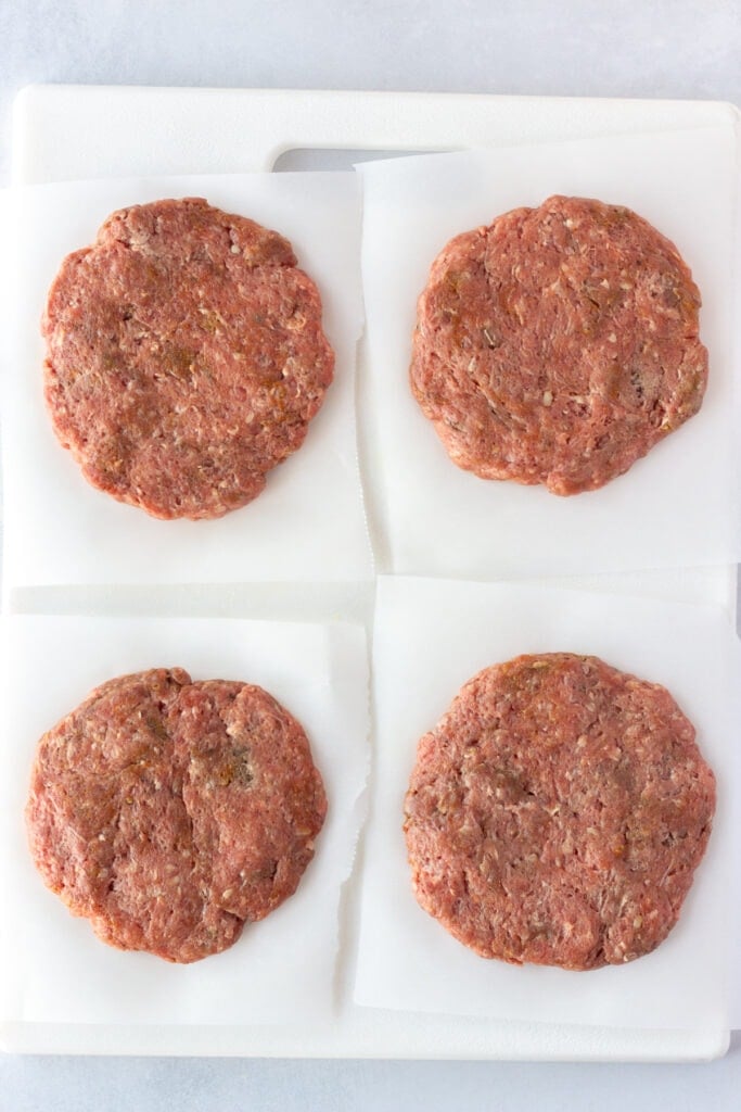 Top down shot of four raw burger patties on top of white parchment paper on a cutting board.