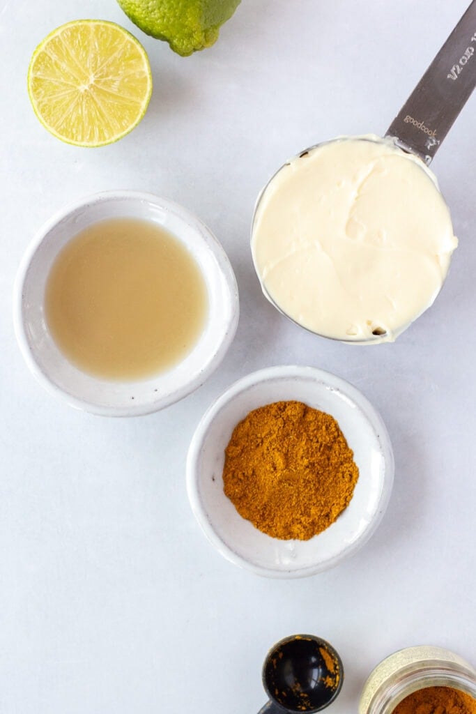 A top down shot of three small containers. One is a silver measuring cup with mayo in, it, another is a small bowl of lime juice, and the last is a small bowl with curry powder. One and a half limes and a measuring spoon with a small jar of curry powder are off to the side.