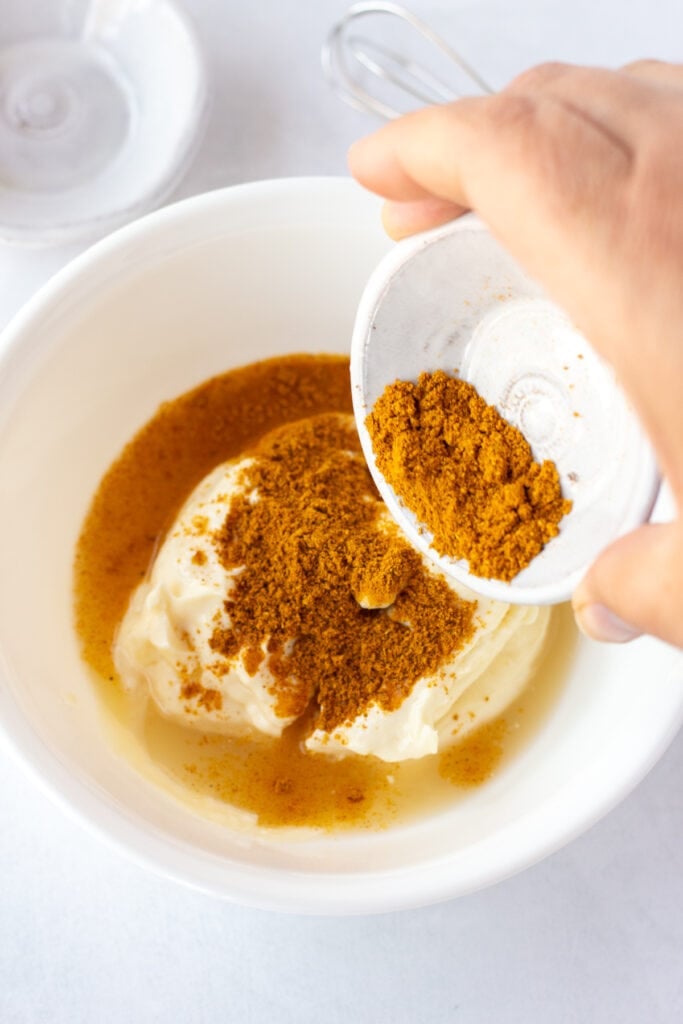 Close up of a hand pouring a small pinch bowl of curry powder into a medium-sized white bowl with mayo and lime juice already in it.