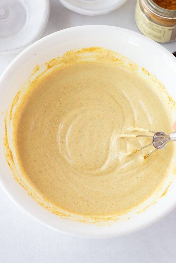 A small whisk gently stirring a curry mayo dip in a medium white bowl that is fully combined.