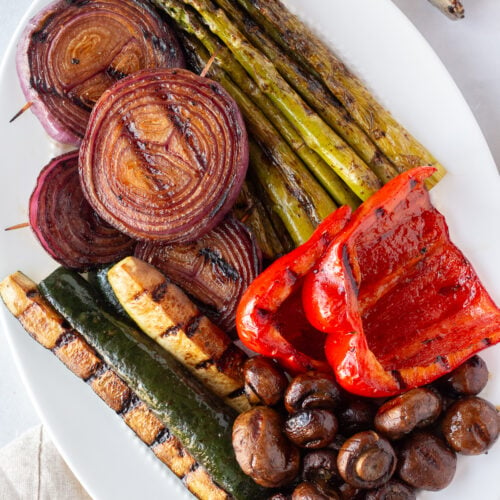 Top down shot of a white oval platter with balsamic grilled veggies on it, including asparagus, red bell pepper, mushrooms, zucchini, and red onion.
