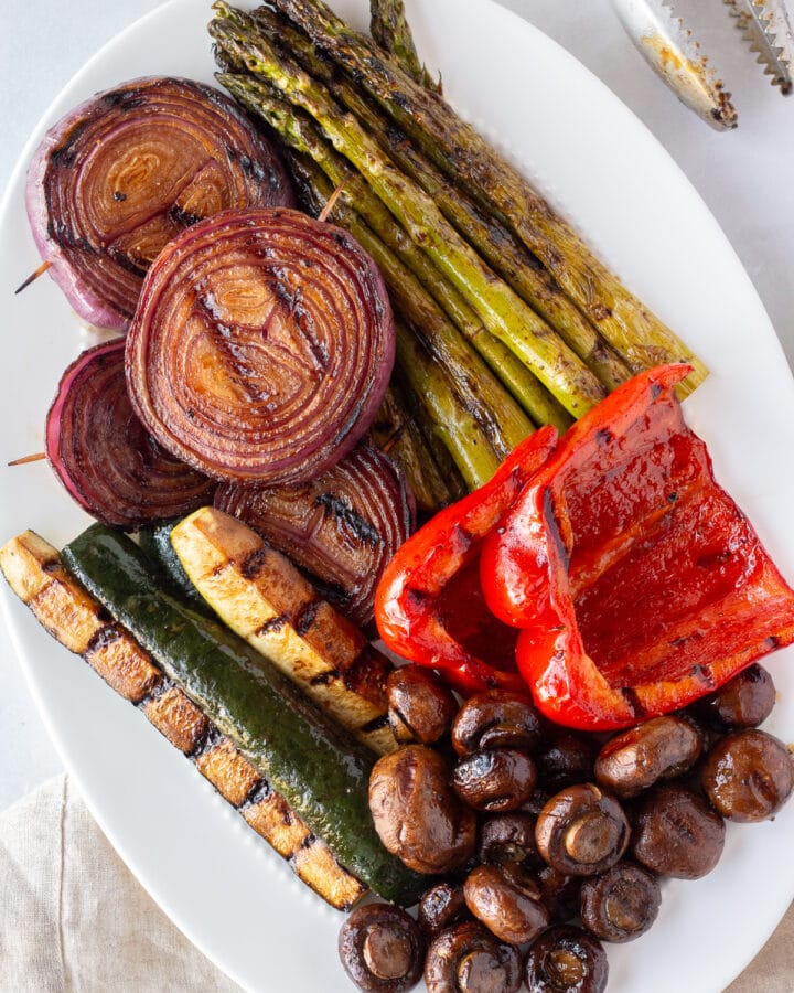 Top down shot of a white oval platter with balsamic grilled veggies on it, including asparagus, red bell pepper, mushrooms, zucchini, and red onion.