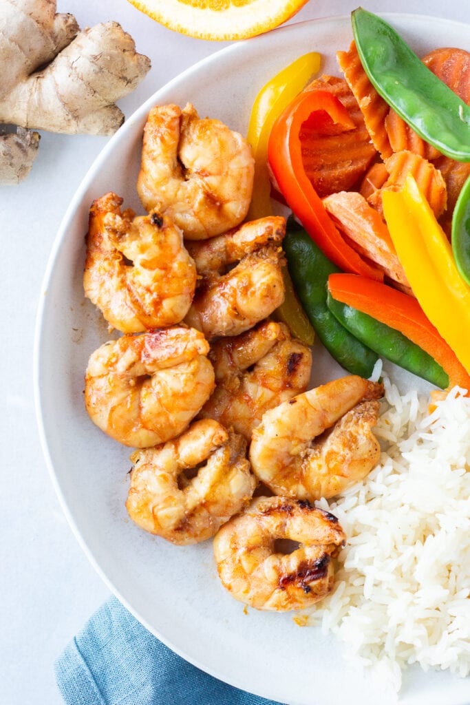 Top down shot of grilled orange shrimp on a white plate with steamed veggies and white rice next to it. A piece or ginger root, a cut orange, and a blue napkin surround the plate.