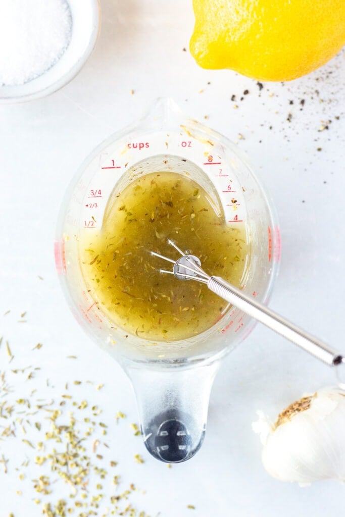 Top down shot of a measuring cup with a lemon herb marinade in it. A small whisk is sticking out of the cup and it rests on a white background, with herbs and a head of garlic surrounding it.