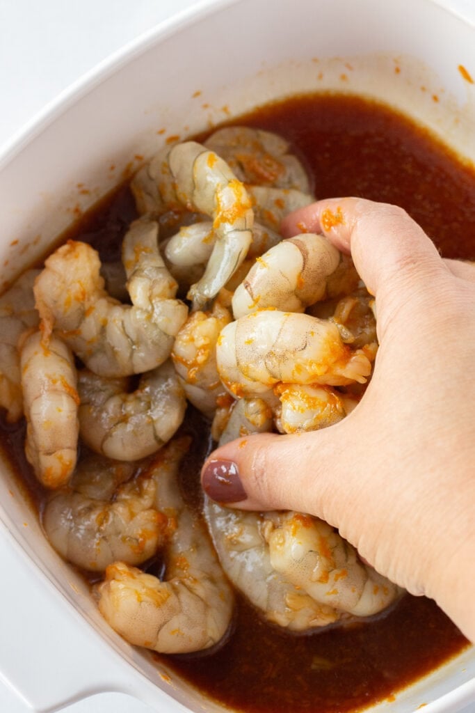 A hand coating raw shrimp in a square white container with an orange soy marinade.