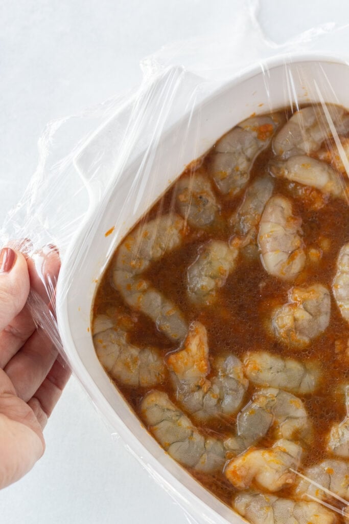 A hand covering a white square container that's filled with raw shrimp in a marinade with clear plastic wrap.