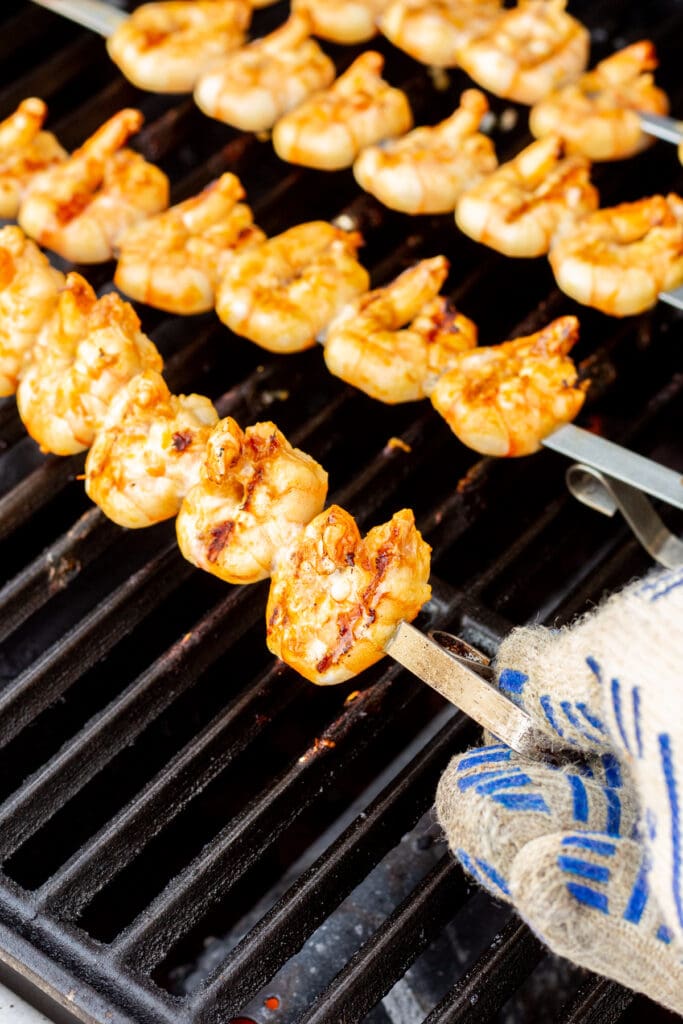 A gloved hand flipping over hot metal shrimp skewers on a grill.